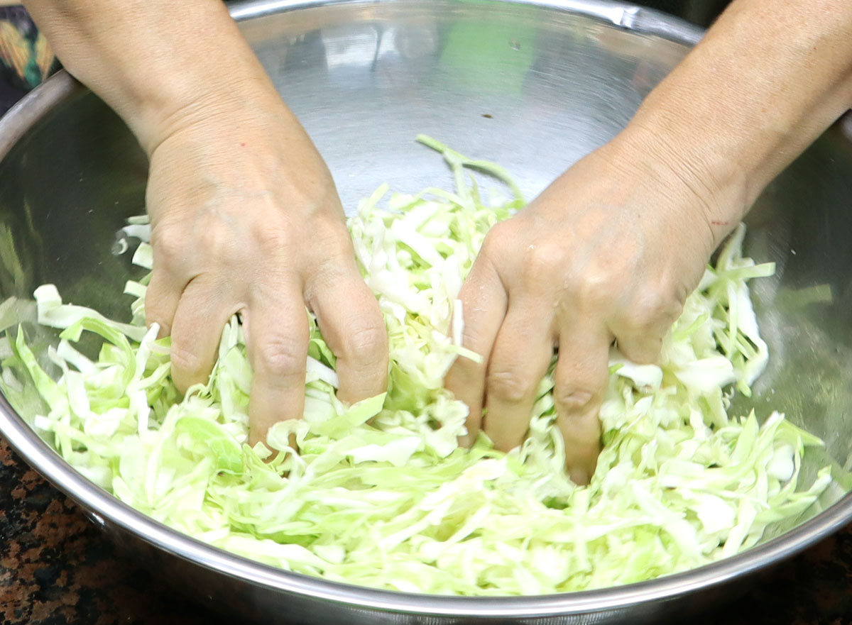 hands crushing cabbage that is in a stainless steel bowl.