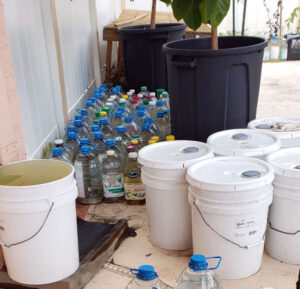 5-gallon buckets and clear plastic jugs of rainwater sitting next to a white fence.