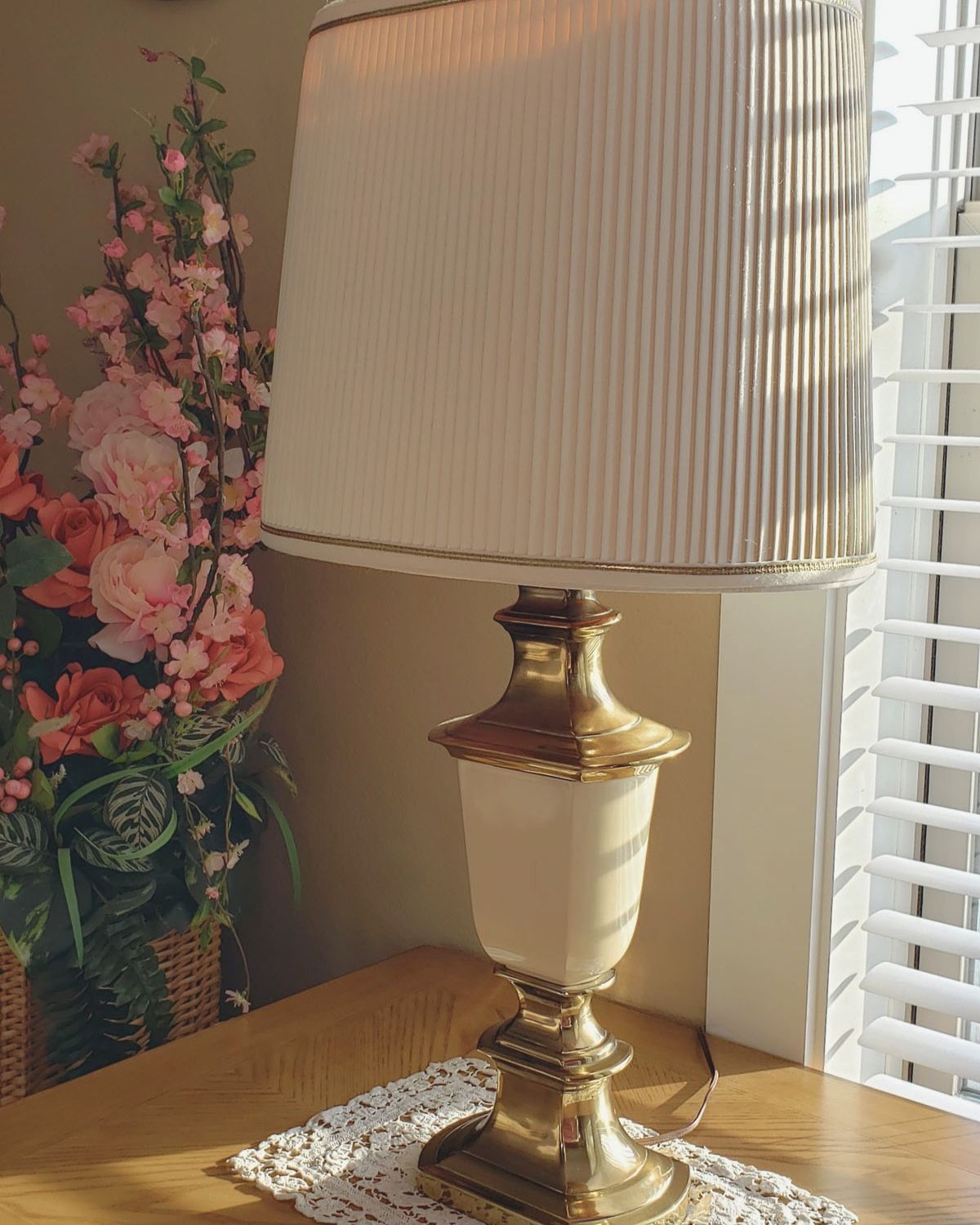 Vintage brass lamp sitting on a woodenn side table next to a window with open blinds letting the sunlight in. An arrangement of red and pink silk flowers is in the background on the left side.