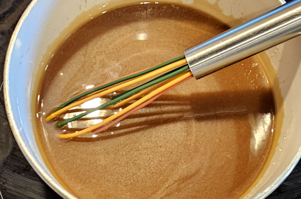 Whisk stirring caramel icing in a small pot on a stove.