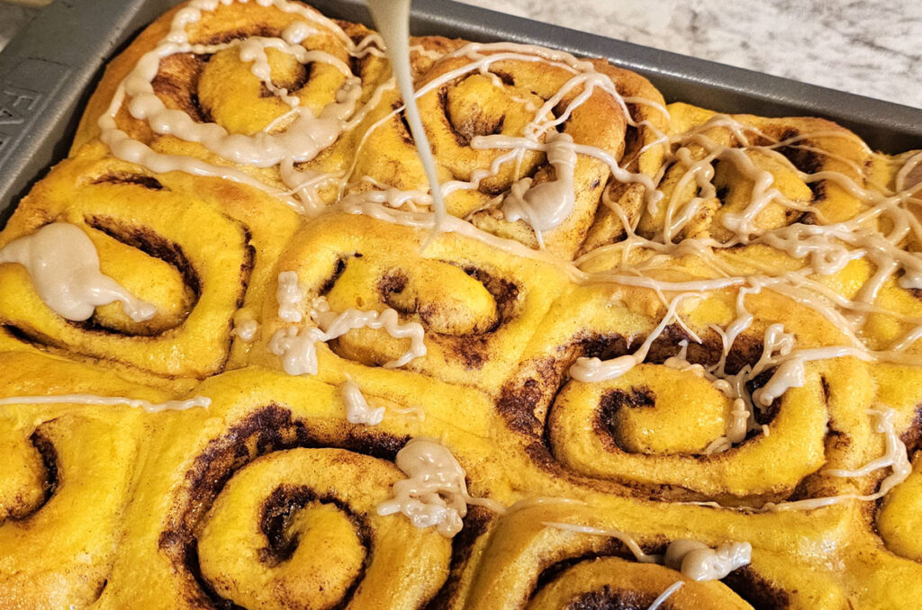 Caramel glaze dripping from a whisk over a pan of freshly baked pumpkin cinnamon rolls.