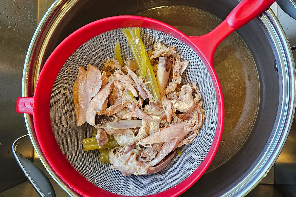 A mesh strainer holding turkey bones and vegetables over a stockpot with turkey broth in it.