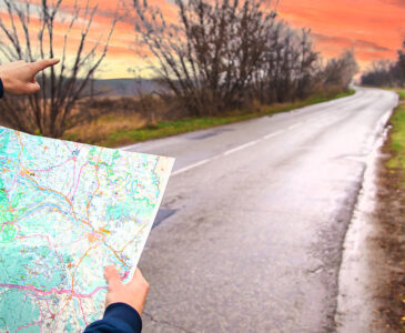 Photo of hands holding a map and pointing down a blacktop road facing a sunset in the countryside.