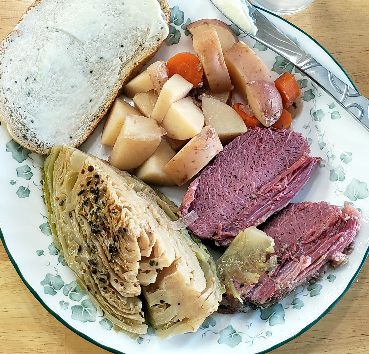 Photo of a plate with corned beef slices, a cabbage wedge sprinkled with spices, carrots and potatoes. A slice of buttered rye bread sits along the edge of the plate.