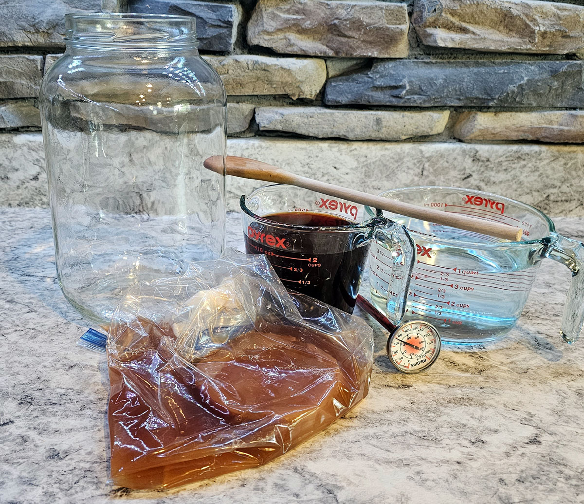 SCOBY in a bag with it's culture tea, a gallon glass jar, glass measuring cup with brewed tea, and a thermometer. Some of the tools for brewing kombucha.