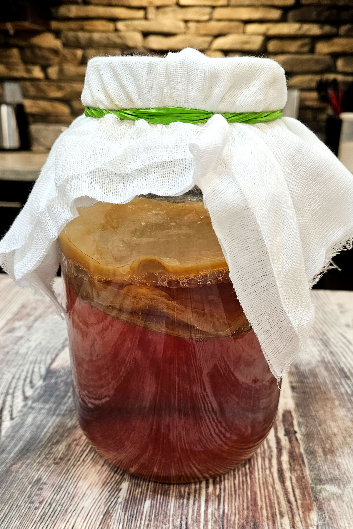 Glass gallon jar of kombucha  on a table. The jar is covered with cheescloth wrapped with a rubber band.