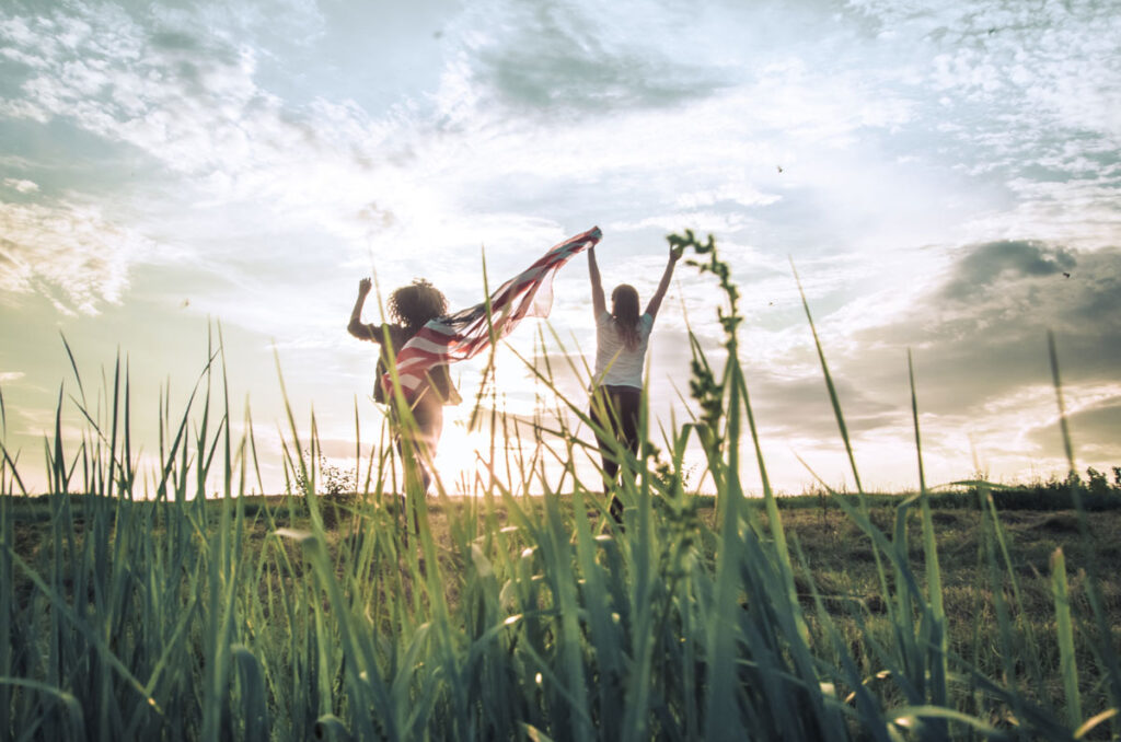 Two young women raising a flag for freedom walking across a wheat field towards the setting sun.