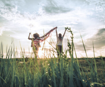 Two young women raising a flag for freedom walking across a wheat field towards the setting sun.