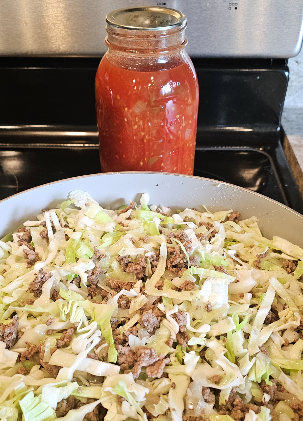 A quart jar of stewed tomatoes on a stovetop next to a frying pan filled with shredded cabbage and browned ground beef.