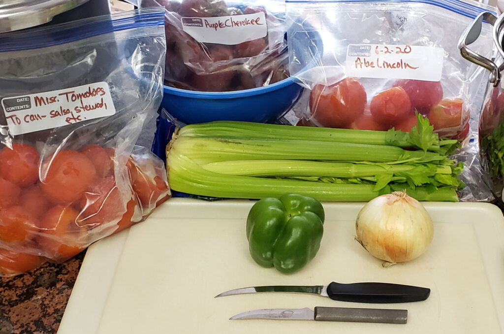 Ingredients for making stewed tomatoes: celery, onion, green pepper, frozen tomatoes. Items are next to a white plastic cutting board with 2 knives on the board.