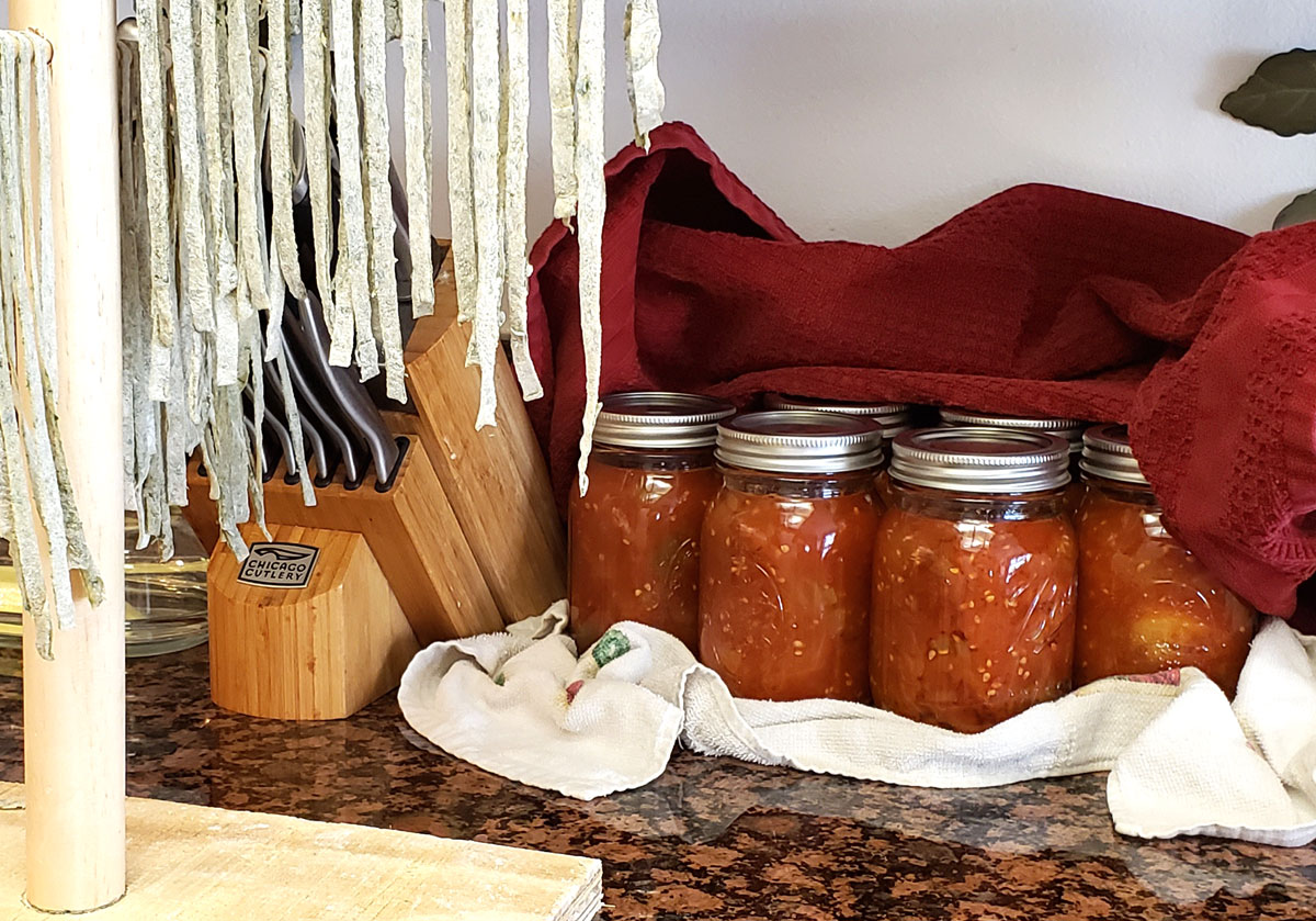 6 pint jars of stewed tomatoes cooling on kitchen towels. They are next to a rack of drying homemade noodles.