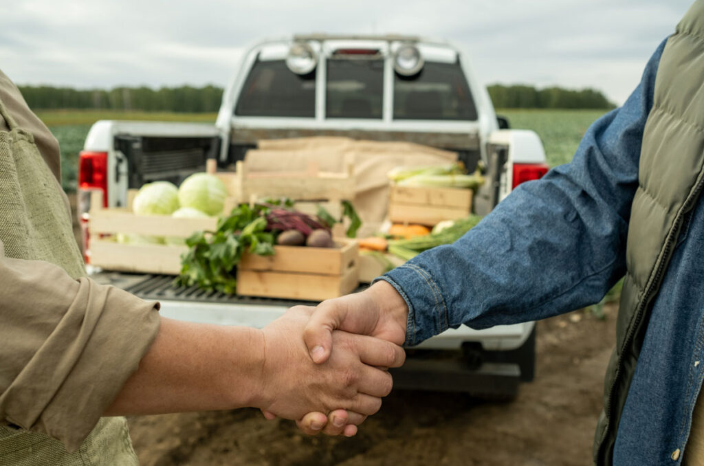 Two growers shaking hands in front of the back of a pickup truck full of fresh vegetables.