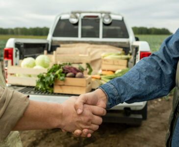 Two growers shaking hands in front of the back of a pickup truck full of fresh vegetables.