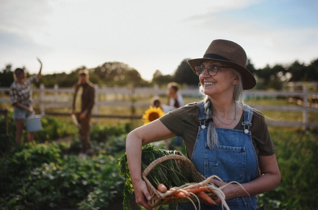 Photo of a woman smiliing while standing in a garden with a basket of produce. Friends are standing and waving to her in the background.