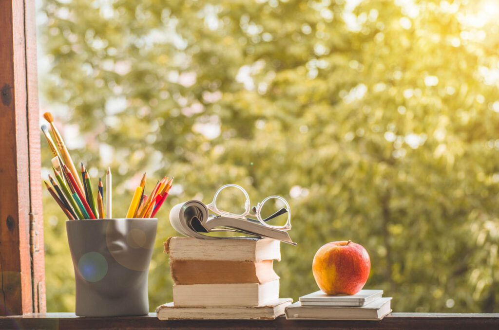 Books, writing tools, an apple, and a pair of reading glasses sitting on a windowsill that faces trees on a sunny day.