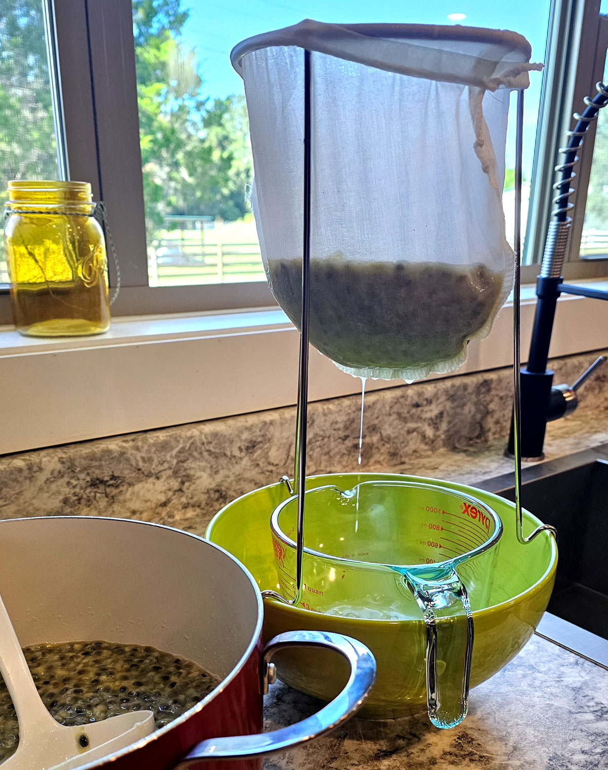 Jelly strainer setup: pot of pulp next to a jelly strainer attached to a large bowl. Juice is dripping from the mesh jelly bag.