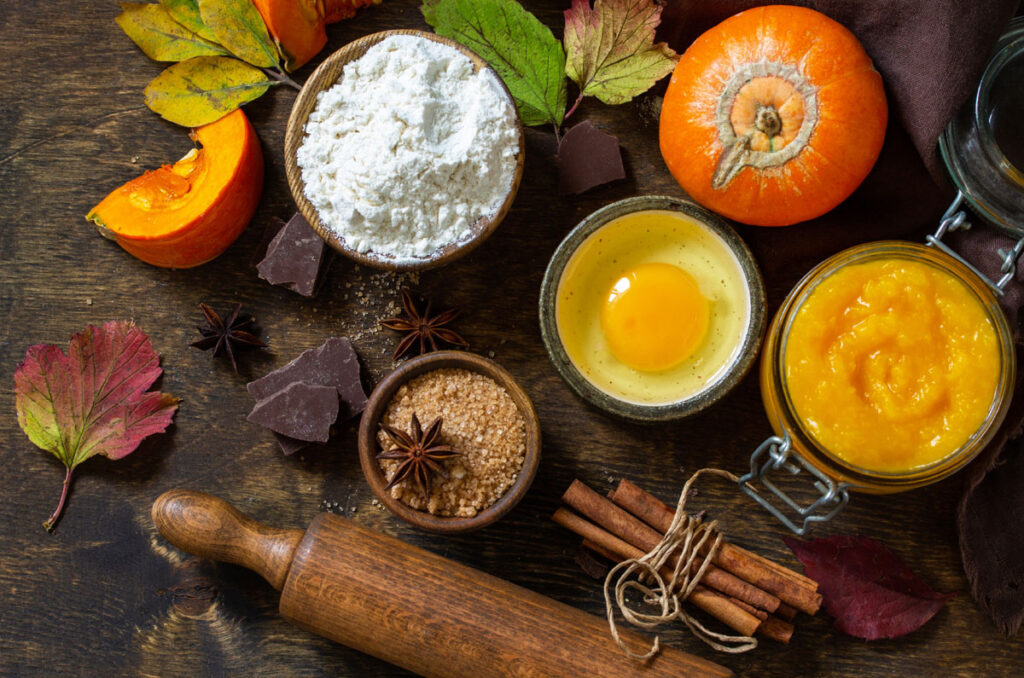 foods and ingredients laid out on a wood table for fall baking: eggs, flour, pumpkin, cinnamon, chocolate pieces, and a wood rolling pin.