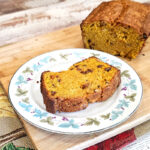 Plate with a slice of butternut squash bread on it. Sitting on a wooden cutting board with cut loaf of bread in the background.