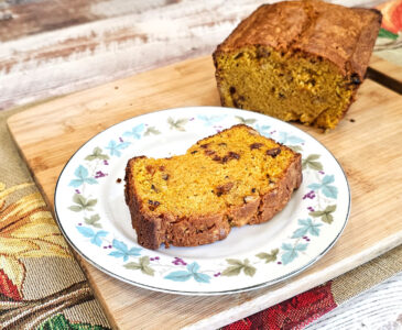 Plate with a slice of butternut squash bread on it. Sitting on a wooden cutting board with cut loaf of bread in the background.