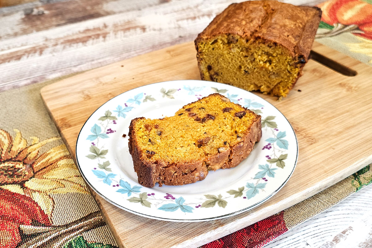 Plate with a slice of butternut squash bread on it. Sitting on a wooden cutting board with cut loaf of bread in the background.