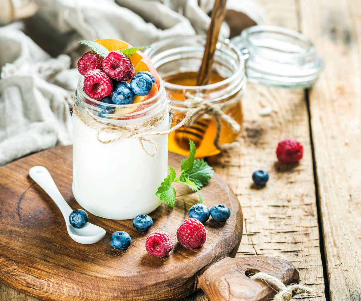 photo of a small glass jar full of yogurt topped with berries with a display of berries on a cutting board, honey, and spoon