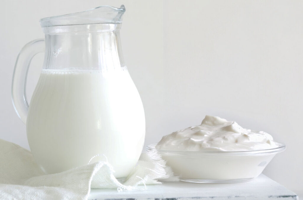 A glass pitcher of milk next to a glass bowl with yogurt, all sitting on a white table top and white background.