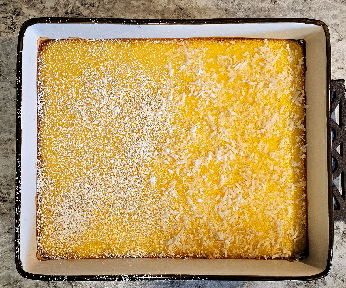 Fully baked papaya bars in baking pan. Left half has sifted powdered sugar on top, right half has coconut flakes sprinkled on top.