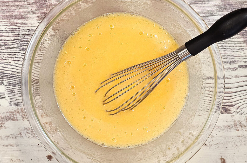 Large glass mixing bowl on a countertop holding whisked eggs, sugar, papaya puree and fruit juices.