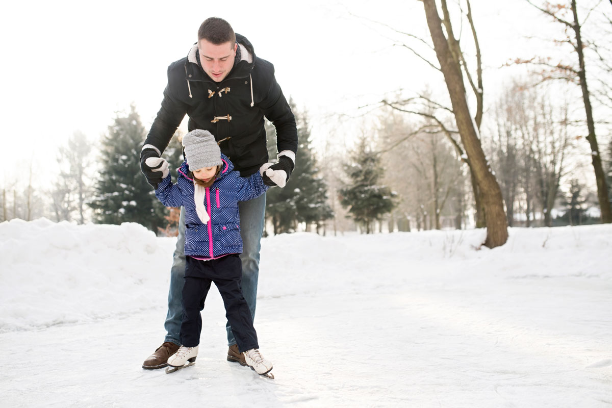 Father and daughter learning to ice skate on a frozen pond near the woods.