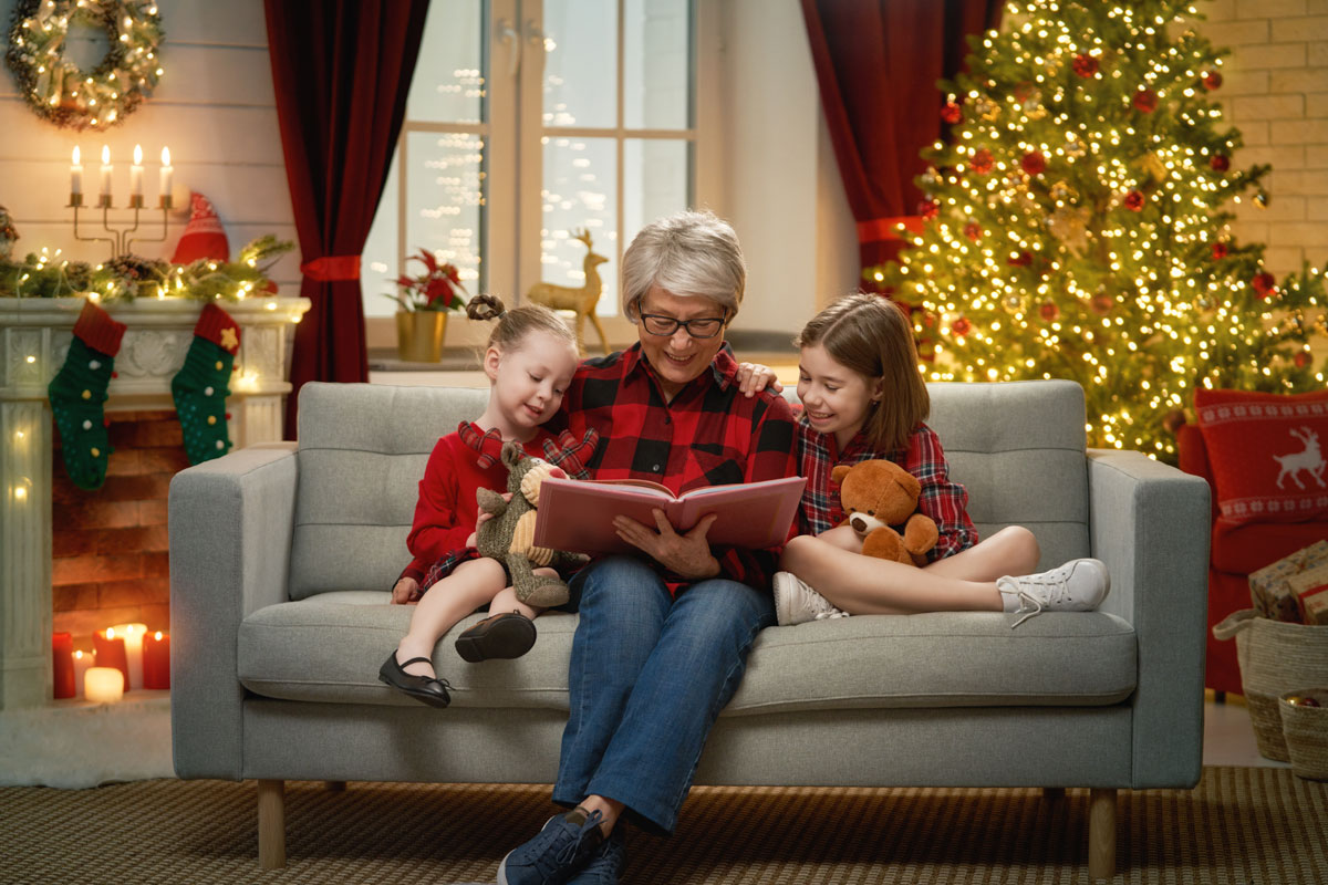 Photo of a grandmother reading books to two grandchildren. They're sitting on a sofa with fireplace and decorated Christmas tree in the background.