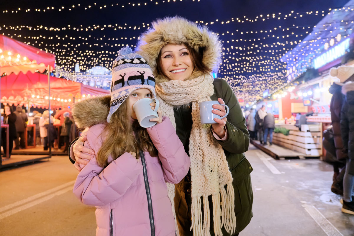 Photo of a mother and daughter at a Christmas festival at night. They're bundled up in coats and holding hot cocoa.