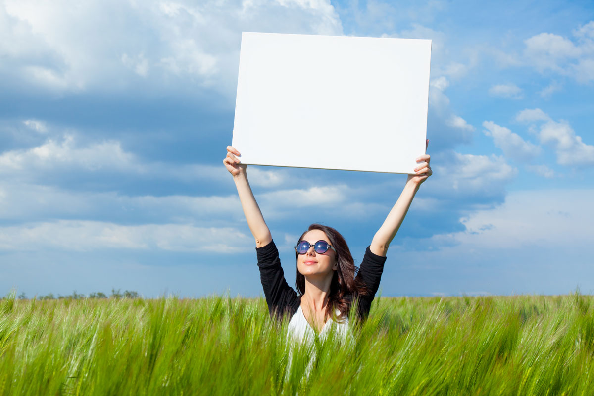 Woman standing in a grassy field holding a blank vision board over her head and wearing sunglasses.