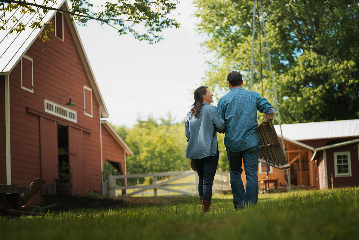 Couple walking across their homestead betwen a barn and outbuilding.