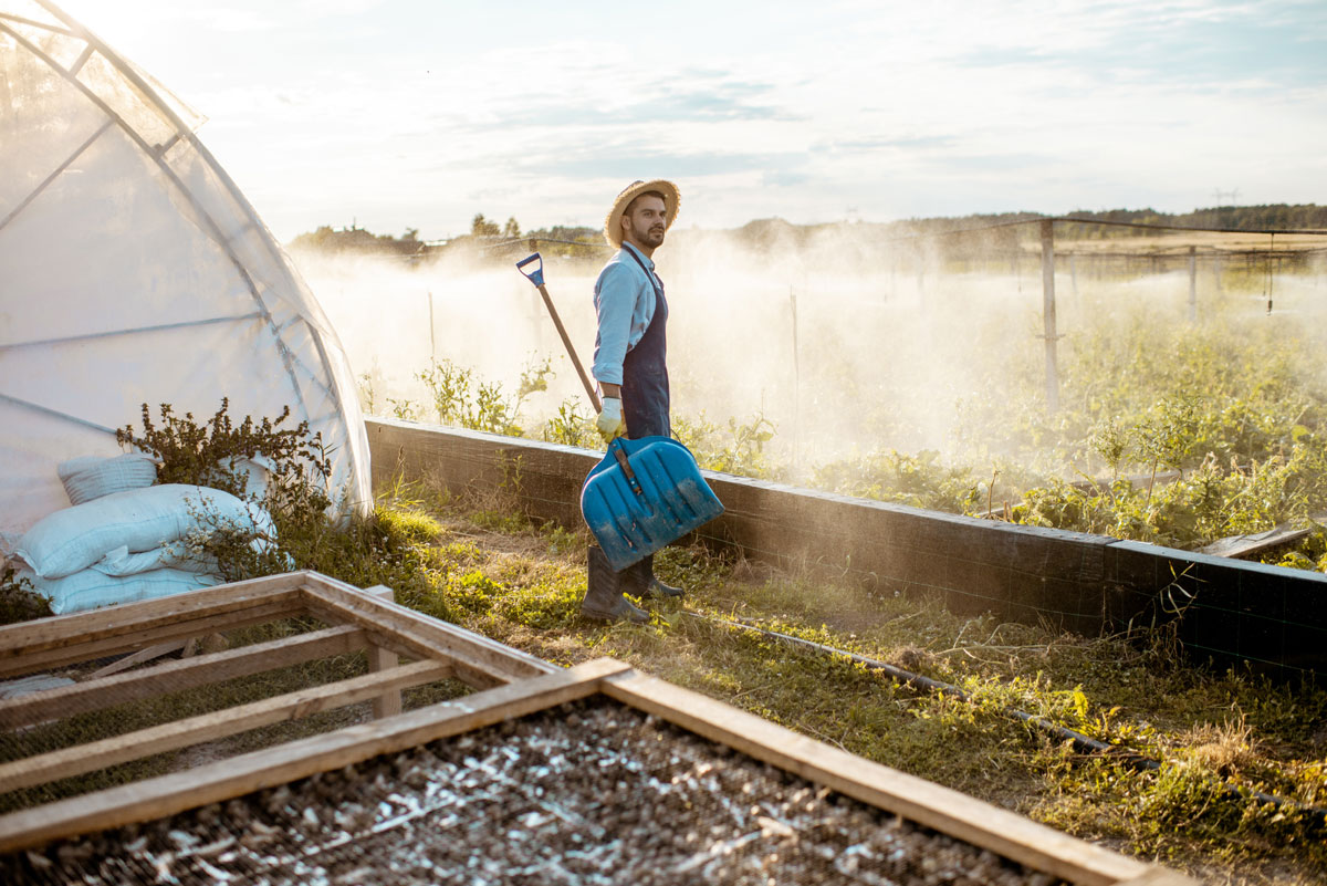 Man standing in front of his greenhouse in the morning sunlight holding a large shovel.