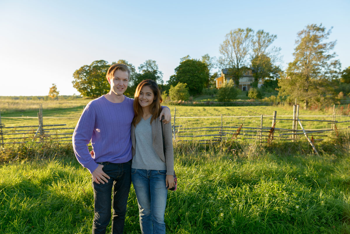 Happy young couple standing in front of their dream homestead. An older farmhouse and fenced pasture are in the background on a sunny summer day.