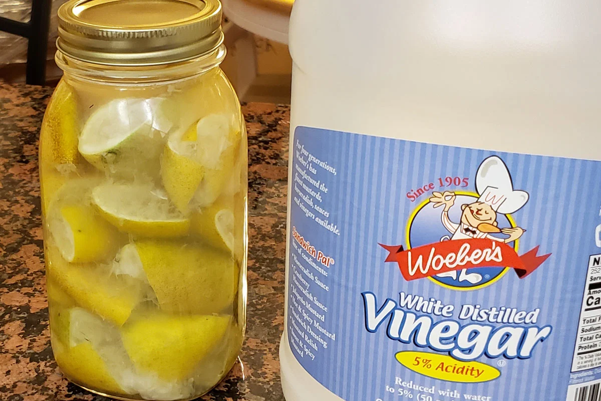 Photo of a quart jar filled with cut up lemons and vinegar next to a container of vinegar on a marble countertop.