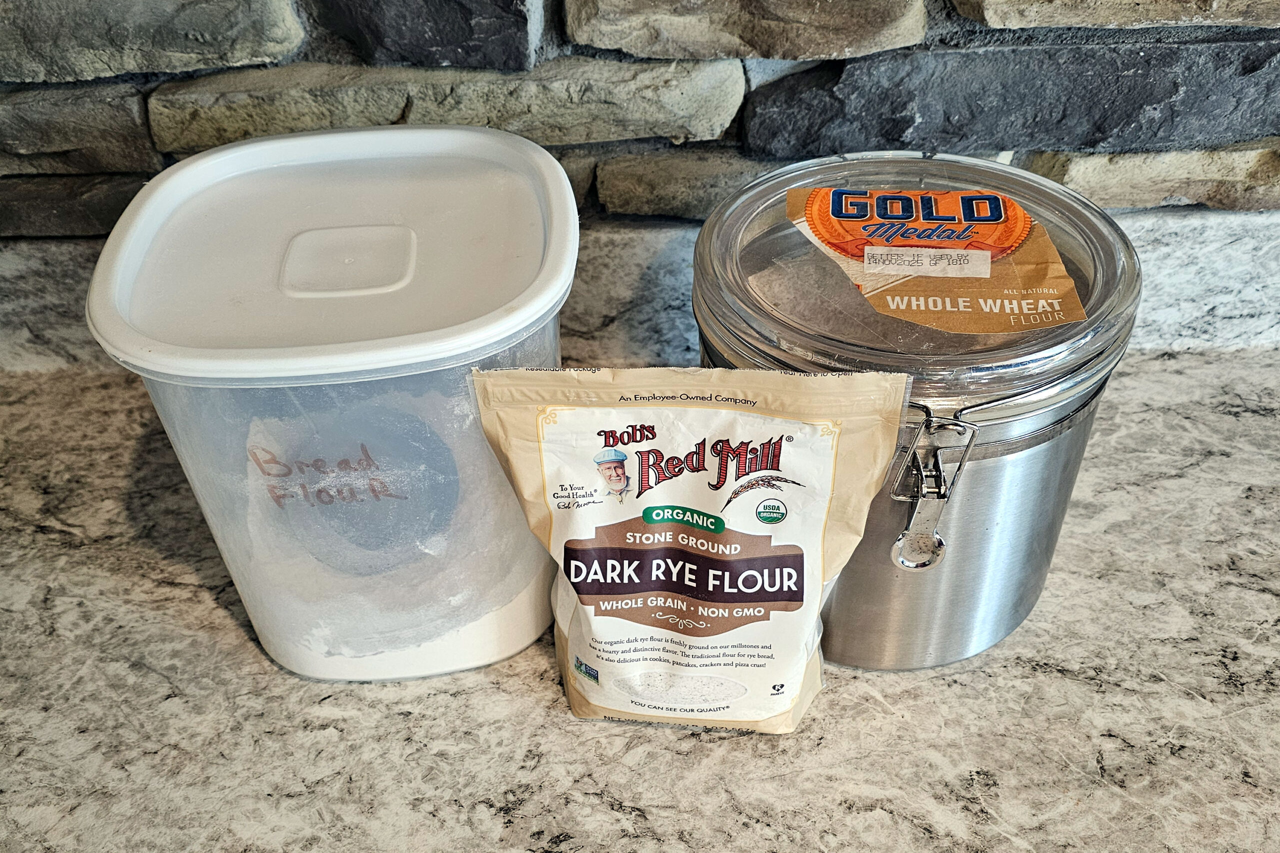 Photo of flours on kitchen counter: unbleached bread flour, rye flour, whole wheat flour.
