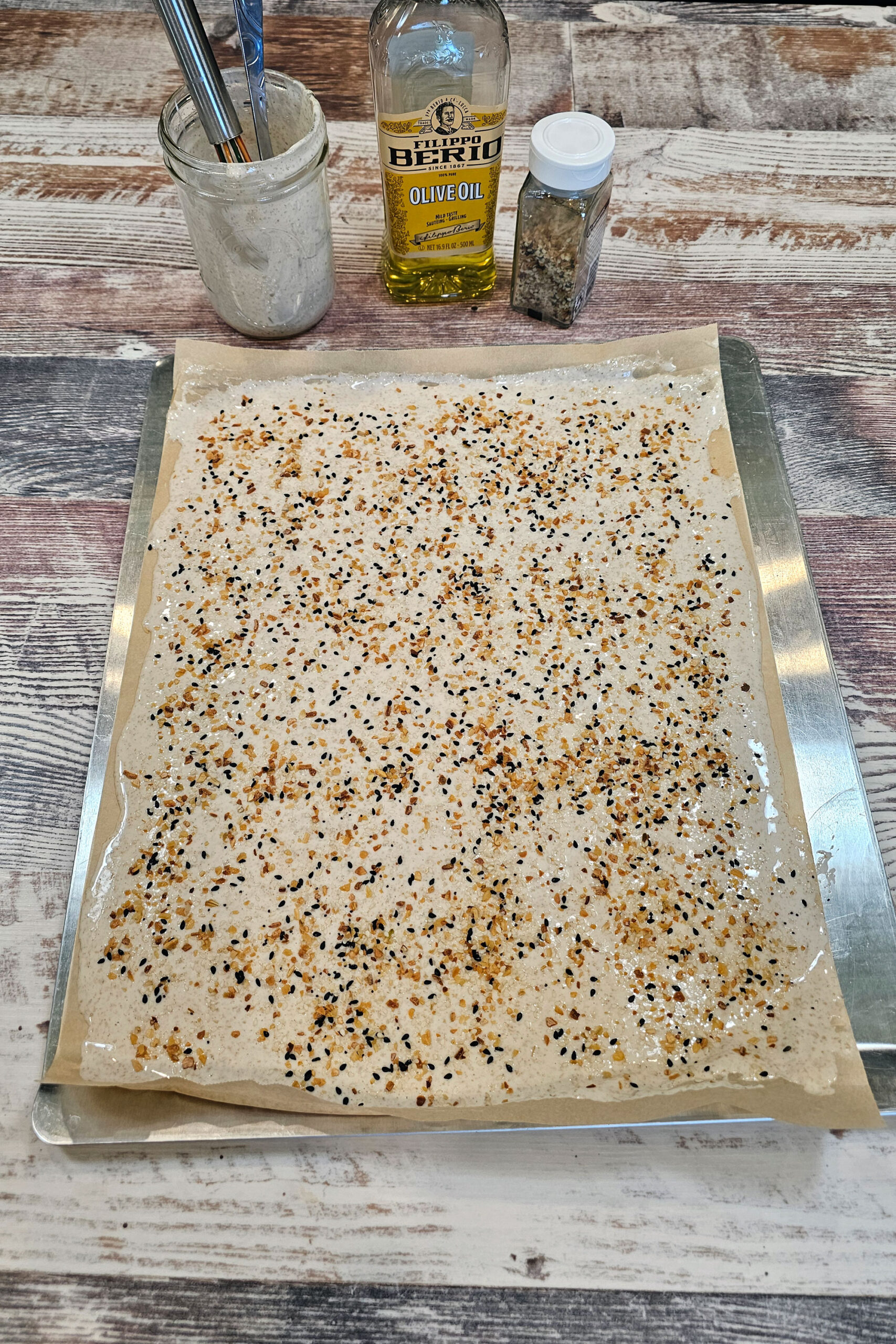 Sourdough discard spread across parchment paper on a baking sheet, sprinkled with seeds, ready to bake.